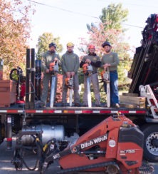 Four workers in safety gear stand on the back of a truck holding chainsaws. Construction equipment, including a Ditch Witch machine, is in front of them—typical of a professional tree service in Montclair NJ. Autumn trees fill the background.