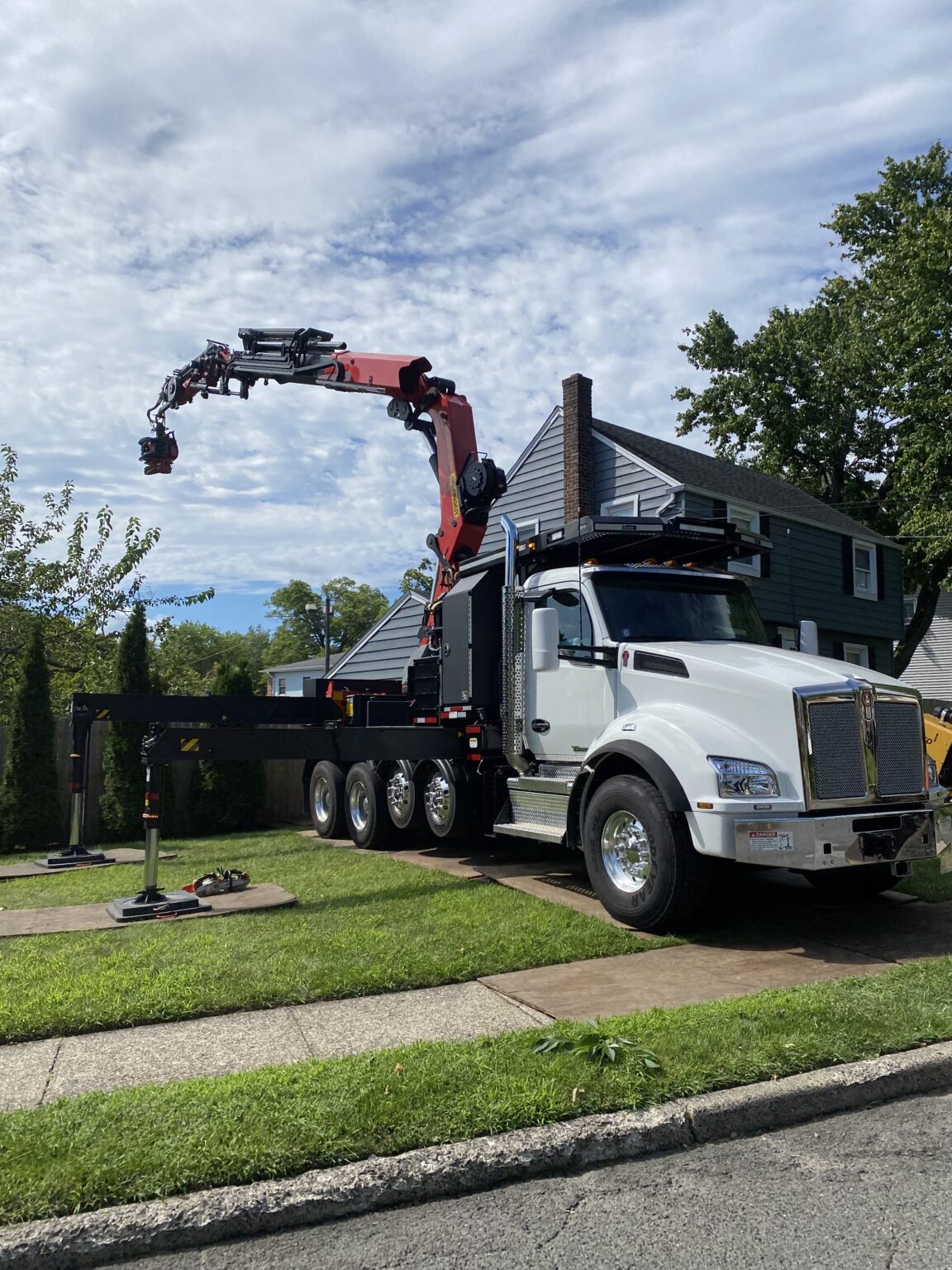 A large white truck with a red hydraulic crane arm, used for tree service in Montclair NJ, is parked on a residential street next to a house, with the crane extended and stabilizers deployed on the grass.