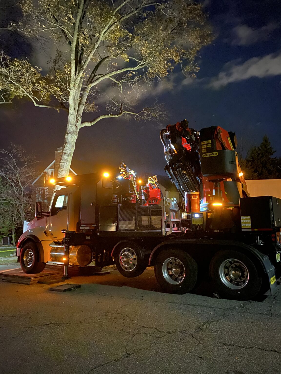 A large truck with a mounted crane, used for tree removal in Montclair NJ, is parked under a tree at night. Its lights illuminate machinery and cast a yellow glow on the surroundings, while clouds and branches are visible in the dark sky above.