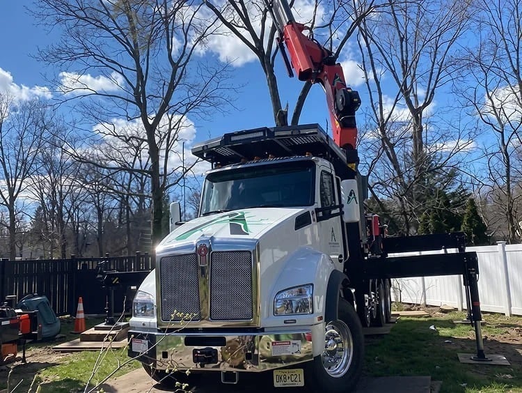 A white truck with a mounted red crane is parked on grass near leafless trees and a black fence under a partly cloudy blue sky.